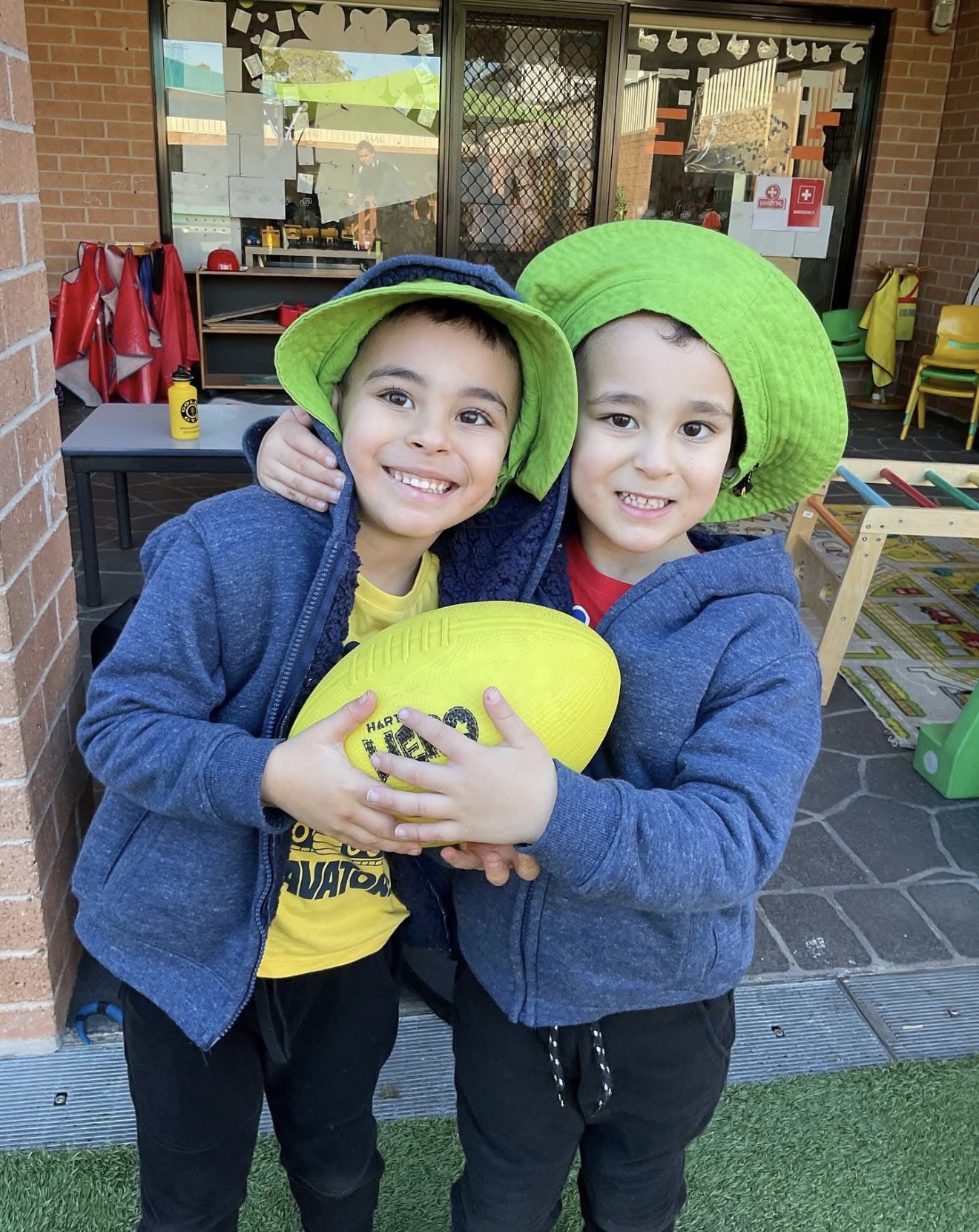Two smiling preschool boys hugging and holding a yellow football during outdoor sports activities at our Punchbowl center