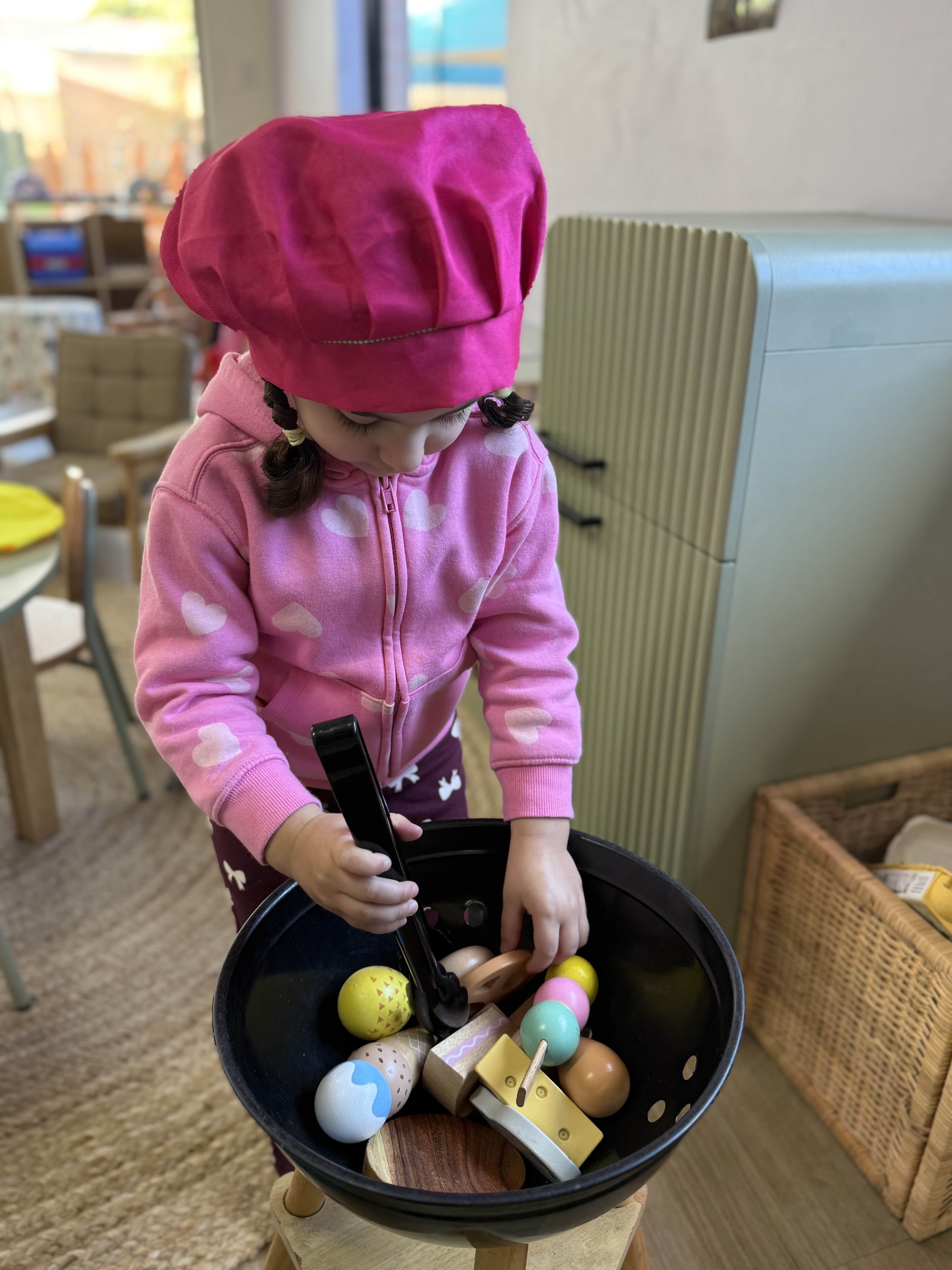 Preschool girl wearing a pink chef hat playing with toy food in a dramatic play kitchen area