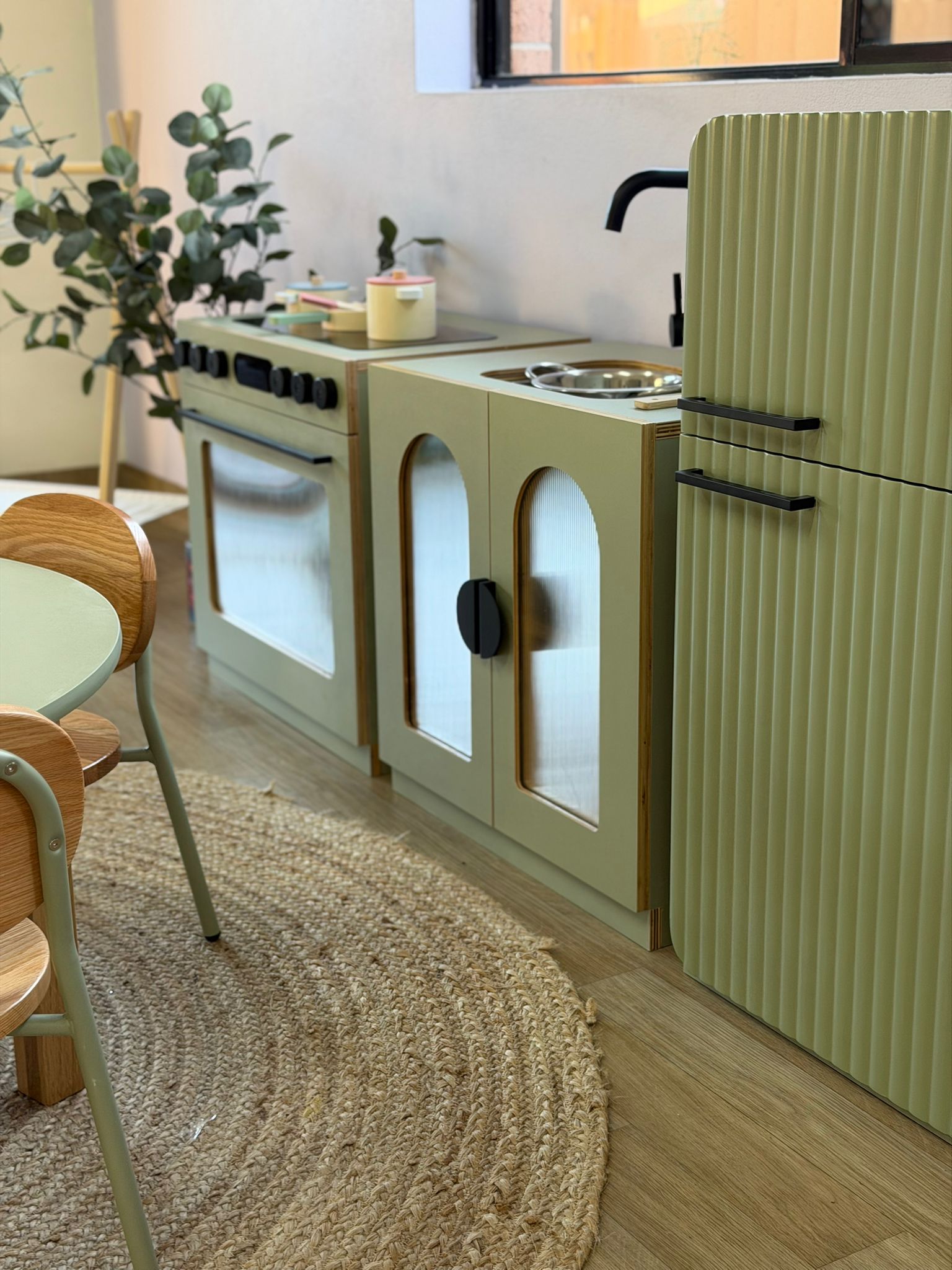 Modern sage green toy kitchen set and a natural woven round rug inside a bright early learning classroom