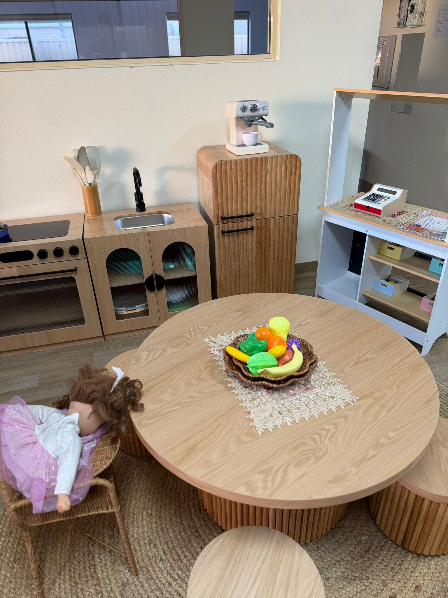 Clean and modern nursery playroom setup featuring a round wooden activity table, doll chair, and play kitchen furniture