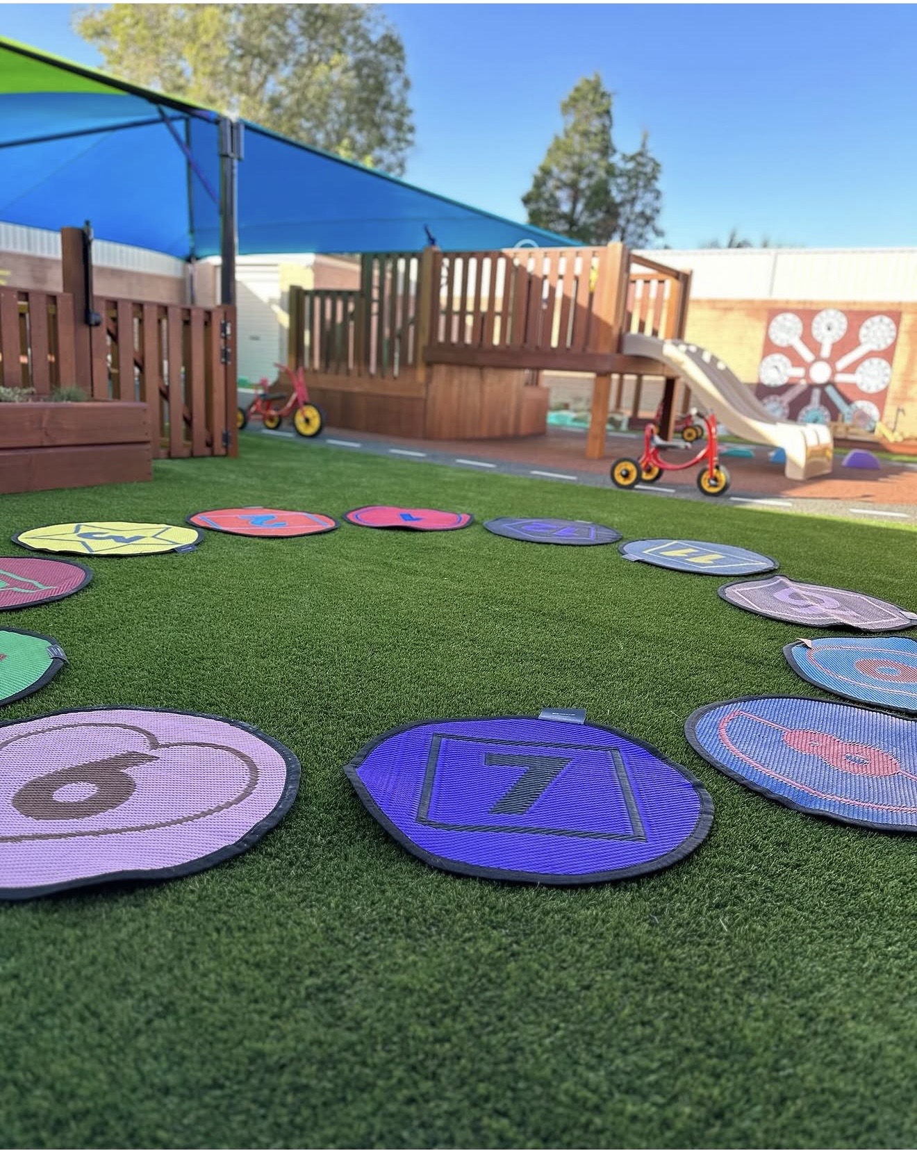 Colorful numbered stepping cushions arranged in a circle on artificial grass in an outdoor preschool playground
