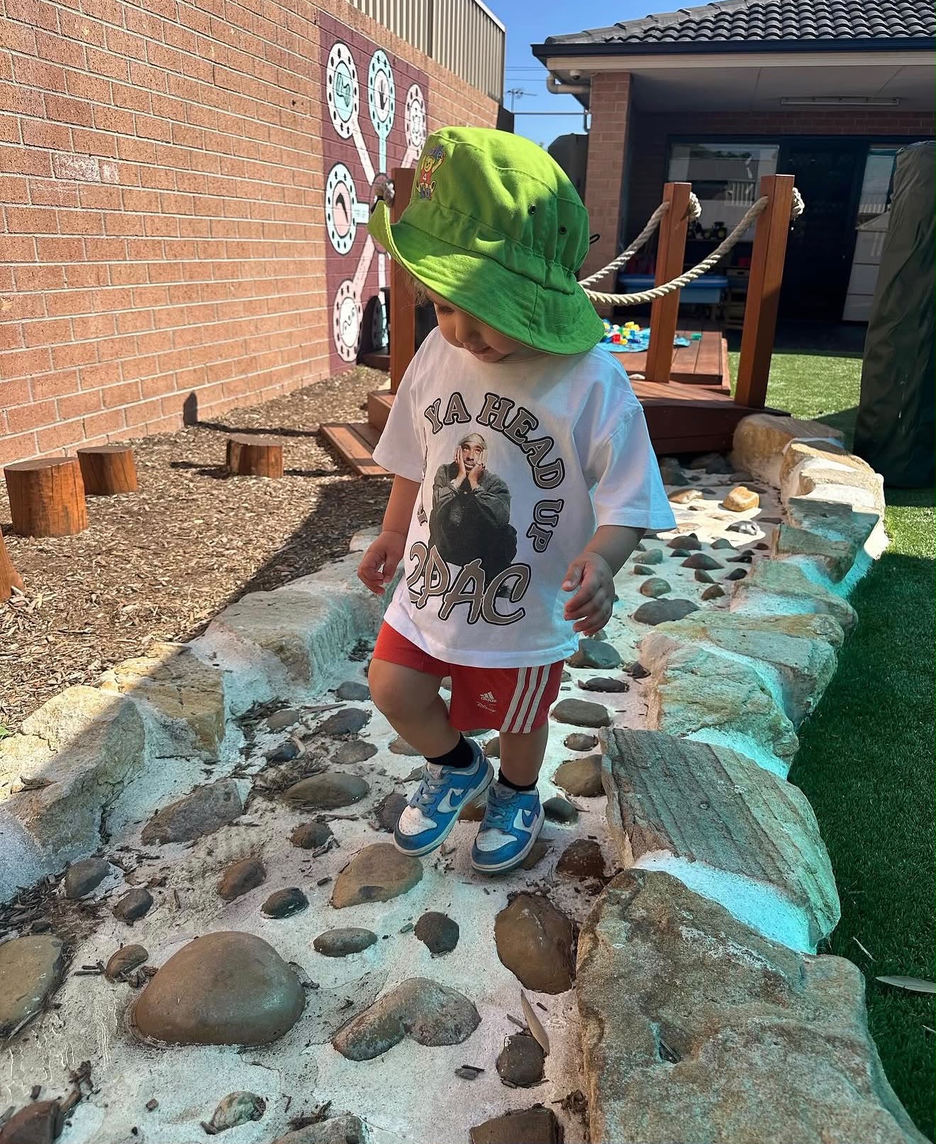 Young toddler wearing a green sun hat carefully navigating an outdoor dry riverbed stone sensory path