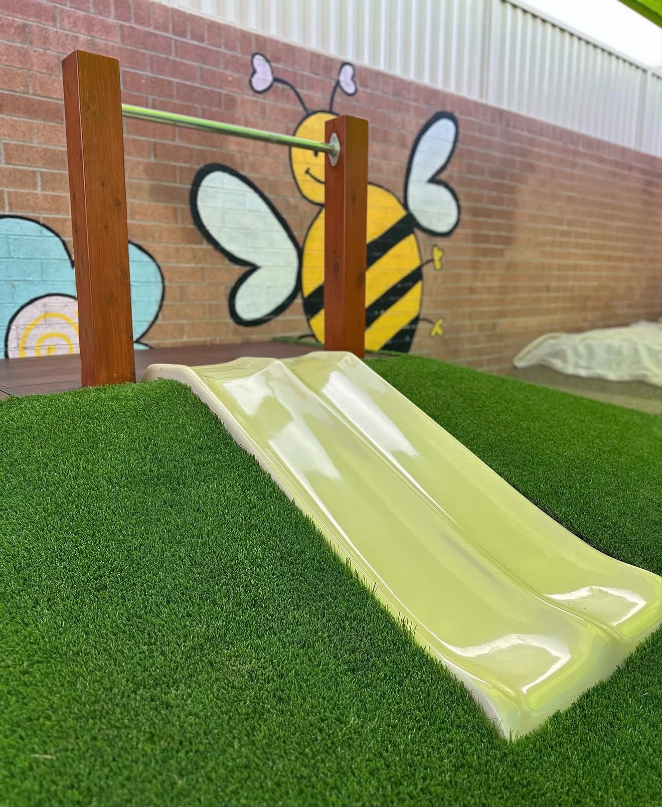 Outdoor playground area featuring a small slide set into a synthetic turf mound under a cheerful bumblebee wall mural