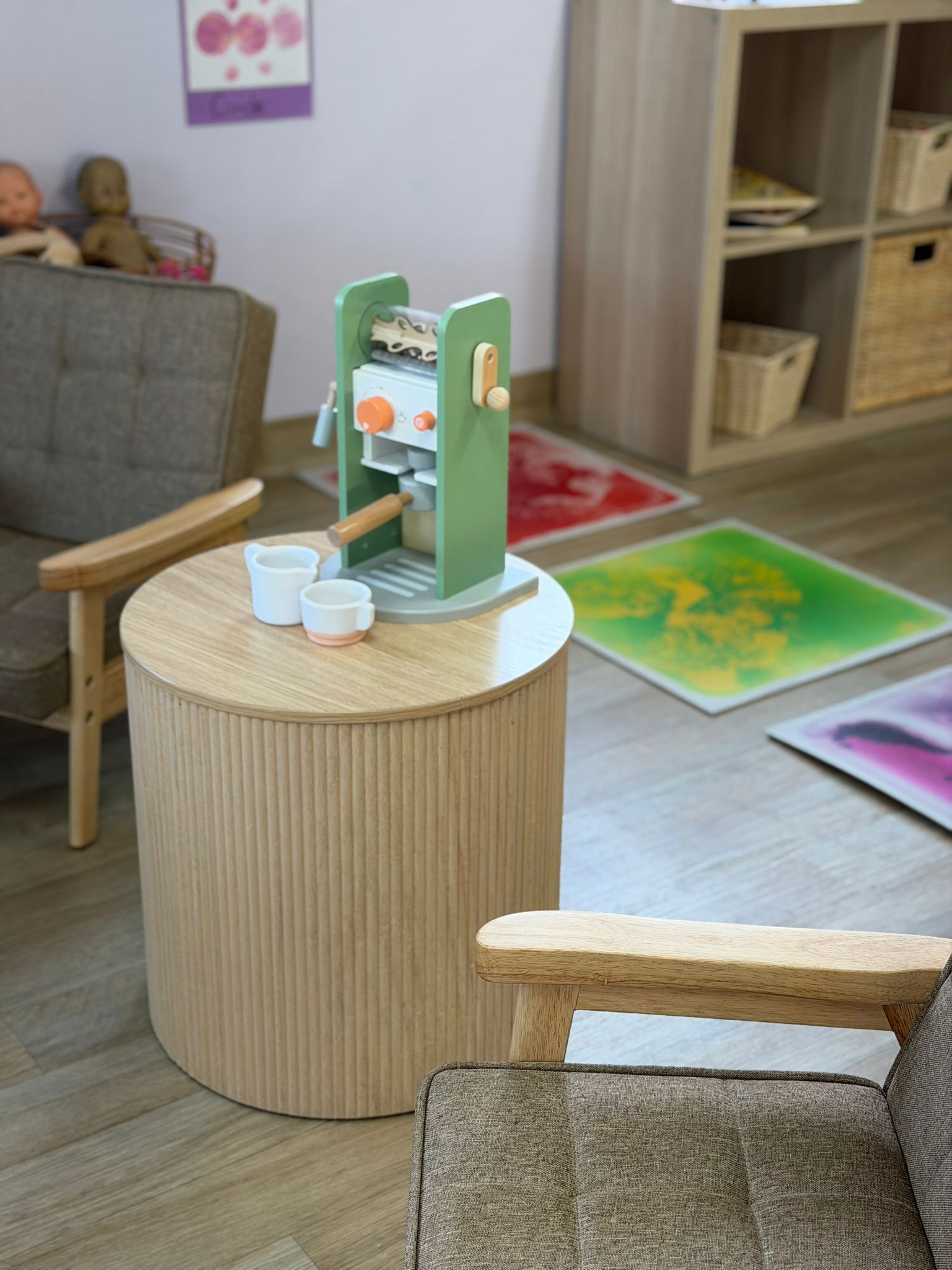 Child-sized wooden espresso machine toy setup on a modern slatted round table next to comfortable armchairs