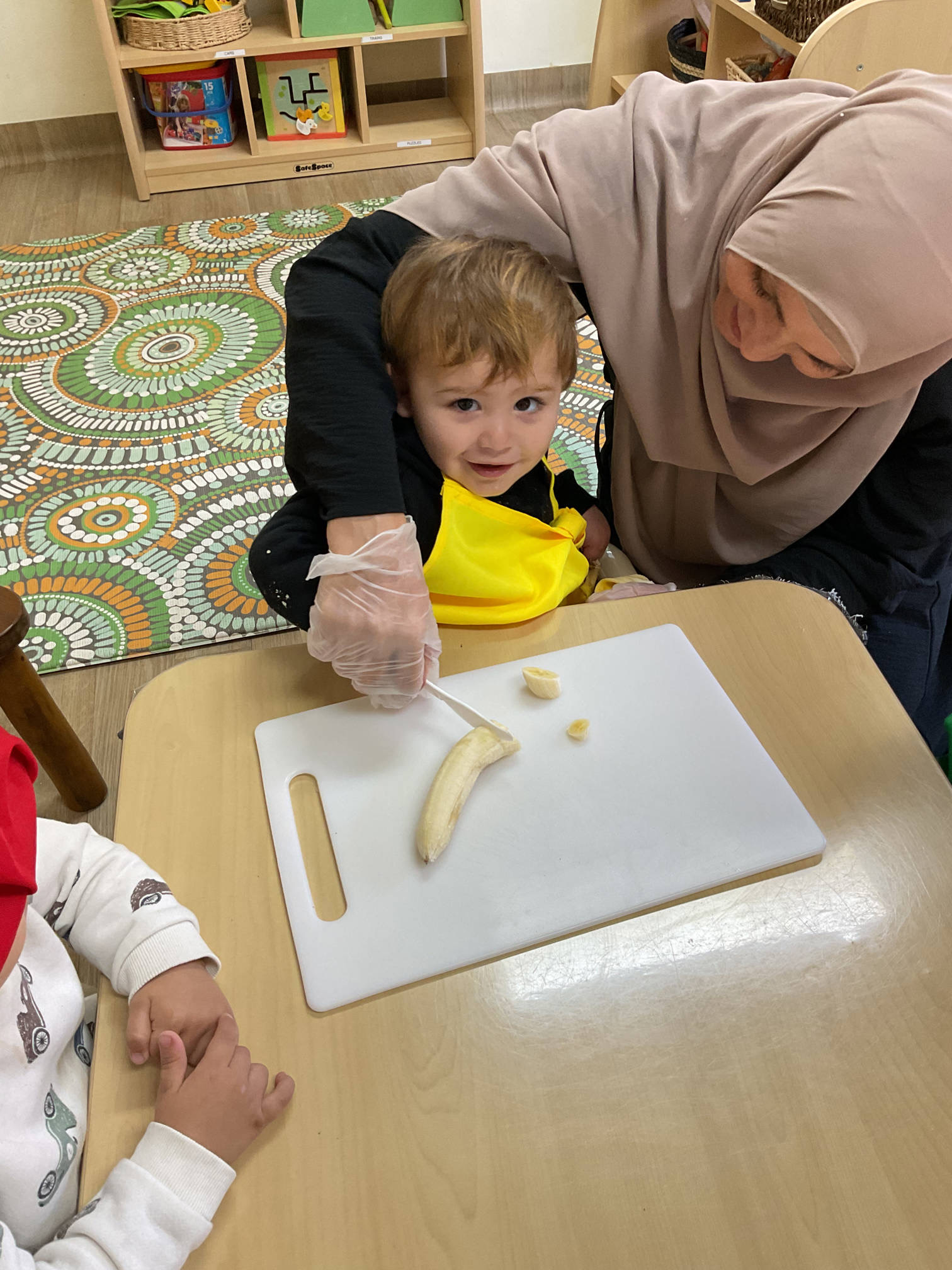 Early childhood educator guiding a young toddler to safely slice a banana during a life skills activity
