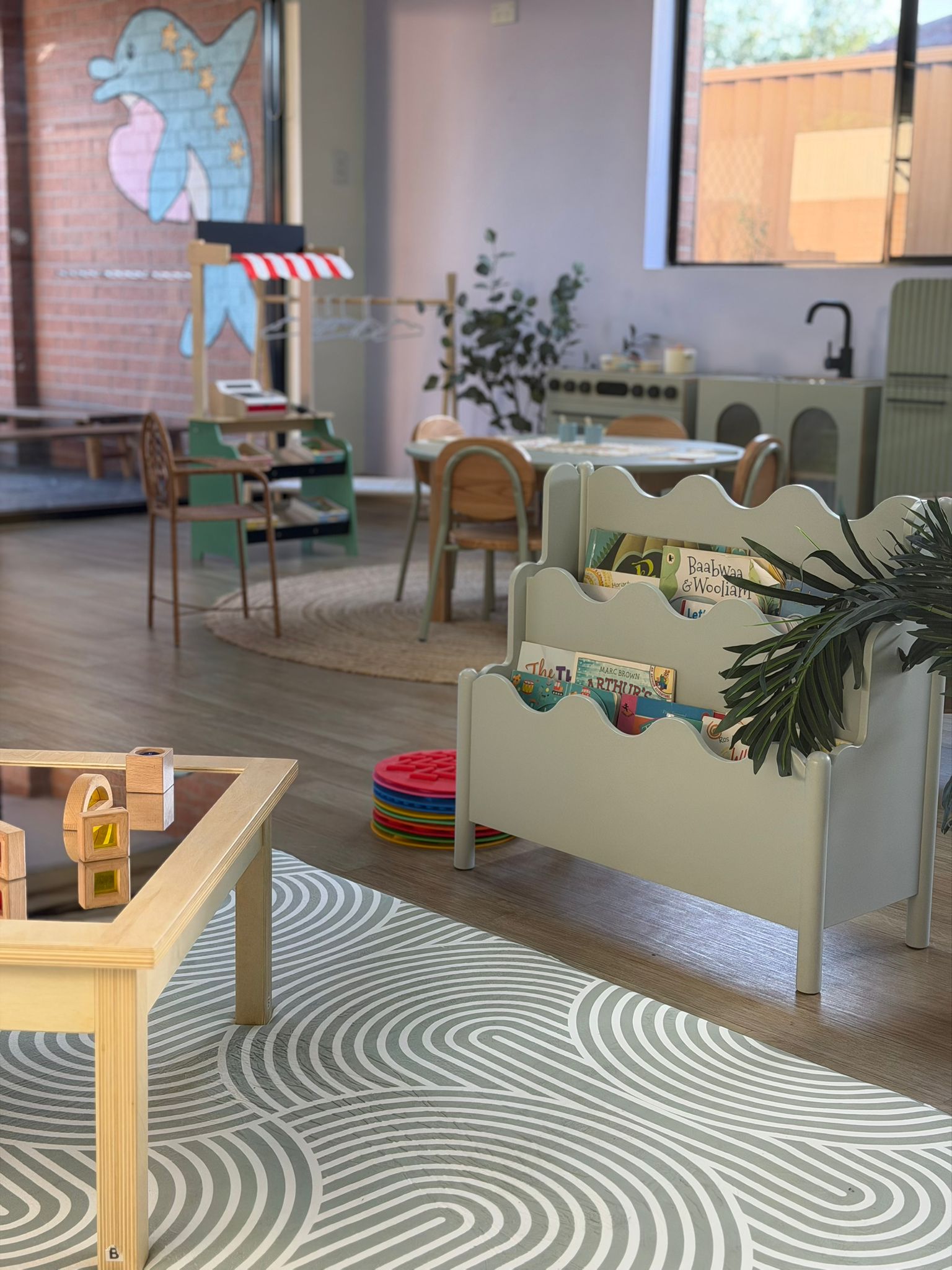 Spacious and sunny open-plan childcare playroom featuring wavy patterned rugs, low tables, and accessible book bins